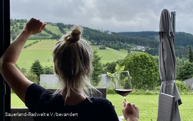 Woman stands with a glass of wine in front of a window showing the green Sauerland landscape.
